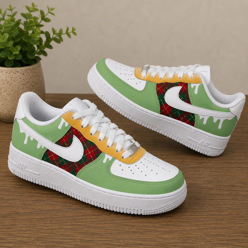 A pair of customized Nike Air Force 1 style sneakers with a green and white color scheme, featuring a red tartan plaid pattern on the Swoosh and heel area, yellow paint-drip accents, and white laces, displayed on a wooden tabletop with a potted plant in the background.