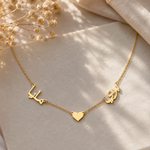 A delicate gold chain necklace featuring three central charms: two Arabic calligraphy characters and a small heart, displayed on a soft beige fabric with dried flowers in the corner.