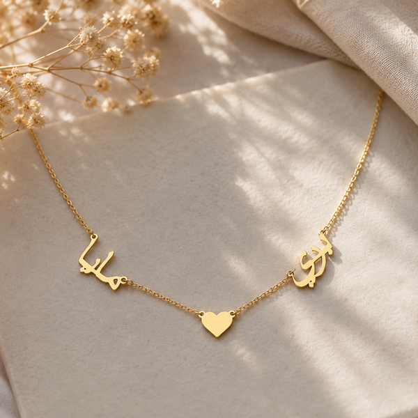 A delicate gold chain necklace featuring three central charms: two Arabic calligraphy characters and a small heart, displayed on a soft beige fabric with dried flowers in the corner.