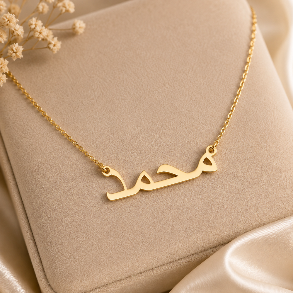 A gold necklace with a horizontal Arabic nameplate pendant, displayed on a beige jewelry box with dried flowers and satin fabric.