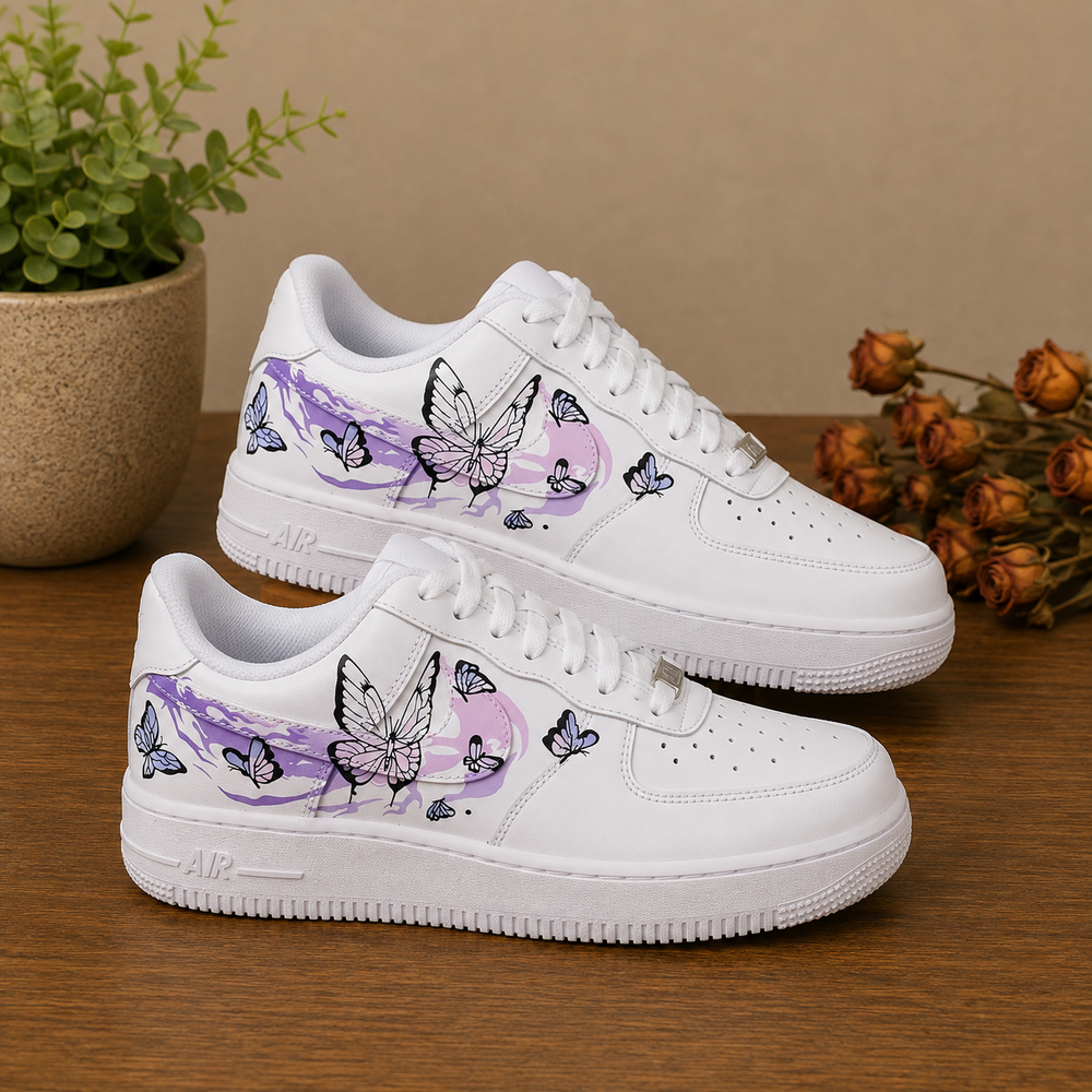 A pair of white low-top sneakers with purple and black butterfly illustrations, displayed on a wooden surface with a potted plant and dried flowers in the background.