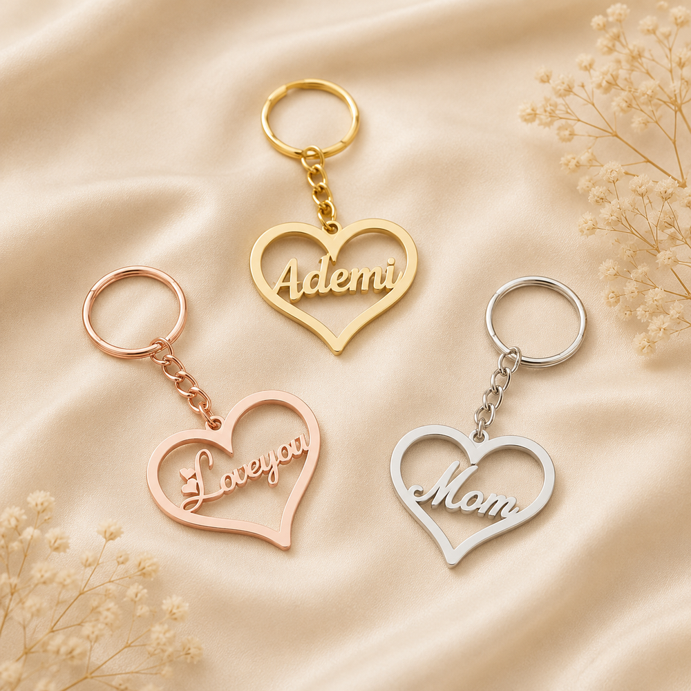 Three heart-shaped keychains with engraved names are arranged on a beige satin fabric background with dried flowers. One keychain is gold with the name 'Ademi', one is rose gold with 'Love you', and one is silver with 'Mom'.
