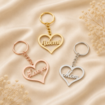 Three heart-shaped keychains with engraved names are arranged on a beige satin fabric background with dried flowers. One keychain is gold with the name 'Ademi', one is rose gold with 'Love you', and one is silver with 'Mom'.