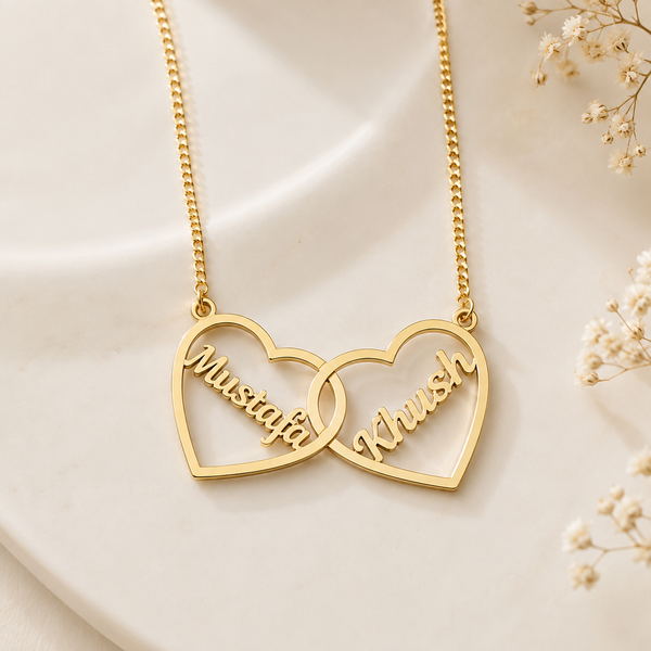 A gold-toned interlocking heart necklace with the names 'Mustafa' and 'Khush' engraved in cursive script, displayed on a soft cream-colored surface with delicate dried flowers in the corner.