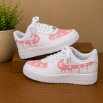 A pair of white low-top sneakers with pink dripping graphic panels and small white bunny motifs, displayed on a wooden surface against a tan background with a potted plant.