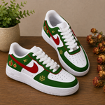 A pair of low-top Nike sneakers with a Christmas holiday theme, featuring green and white panels, red swooshes, gold snowflakes, and festive accents, displayed on a wooden surface with a potted plant and dried flowers.