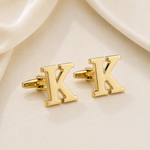 A pair of gold initial cufflinks shaped like the letter 'K', displayed on a soft cream satin fabric background.
