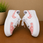 A pair of white low-top sneakers with pink graphic overlays and small white bird illustrations, displayed on a wooden surface against a tan background with a potted plant.