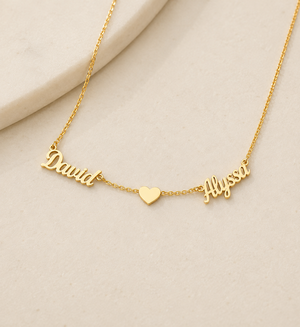 A delicate gold chain necklace featuring personalized nameplates spelling 'David' and 'Hysa' separated by a small heart charm, displayed on a light beige textured surface.
