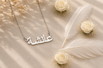 A silver Arabic nameplate necklace with the name 'Alia' (ألياء) in elegant calligraphy, displayed on a beige satin fabric with decorative white feathers and small white roses.