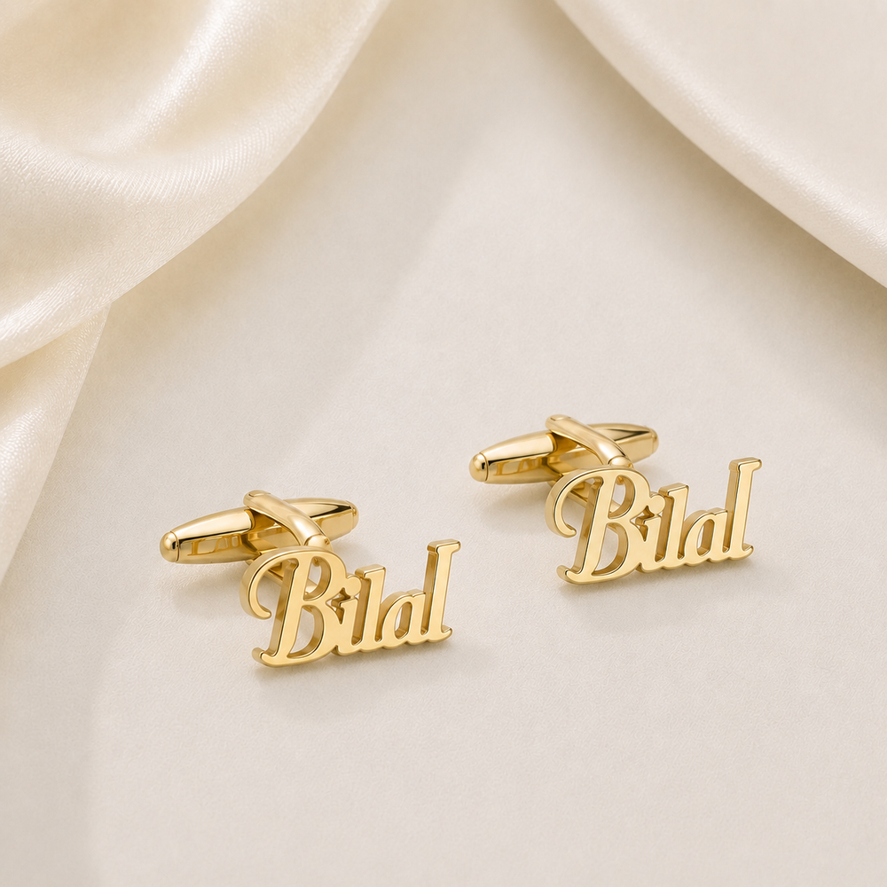 A pair of gold cufflinks shaped like the name 'Bilal' in cursive script, displayed on a soft cream satin background.