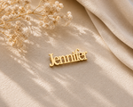 A gold nameplate pin spelling 'Jennifer' in a cursive font, displayed on a textured beige fabric with dried baby's breath flowers and soft shadows.