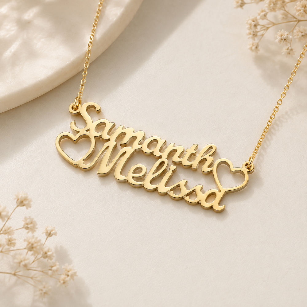 A gold nameplate necklace spelling 'Samanth Melissa' with heart details, displayed on a light cream surface with dried flowers and a marble slice.