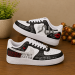 A pair of custom white and black low-top sneakers with graphic prints, displayed on a wooden surface with decorative plants and dried flowers in the background.