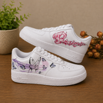 A pair of white low-top sneakers with hand-painted butterfly and floral designs, displayed on a wooden surface with a potted plant and dried flowers in the background.