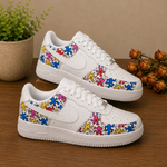 A pair of white Nike Air Force 1-style sneakers decorated with colorful, repeating human figure patterns in pink, blue, yellow, and purple. The shoes are displayed on a wooden table with decorative plants in the background.