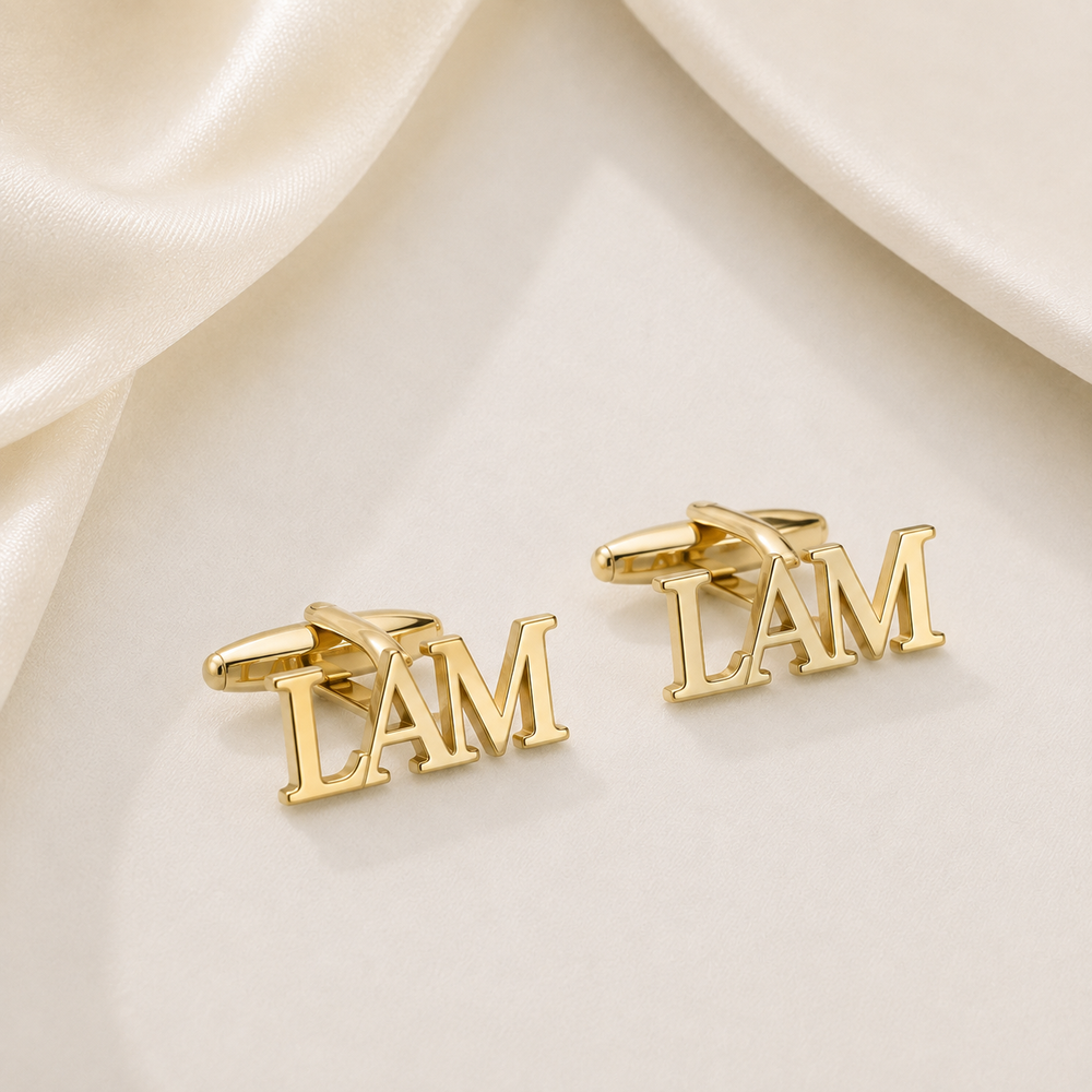 A pair of gold cufflinks shaped like the letters 'I AM' in 3D block lettering, displayed on a cream satin fabric background.