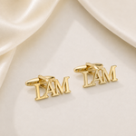 A pair of gold cufflinks shaped like the letters 'I AM' in 3D block lettering, displayed on a cream satin fabric background.
