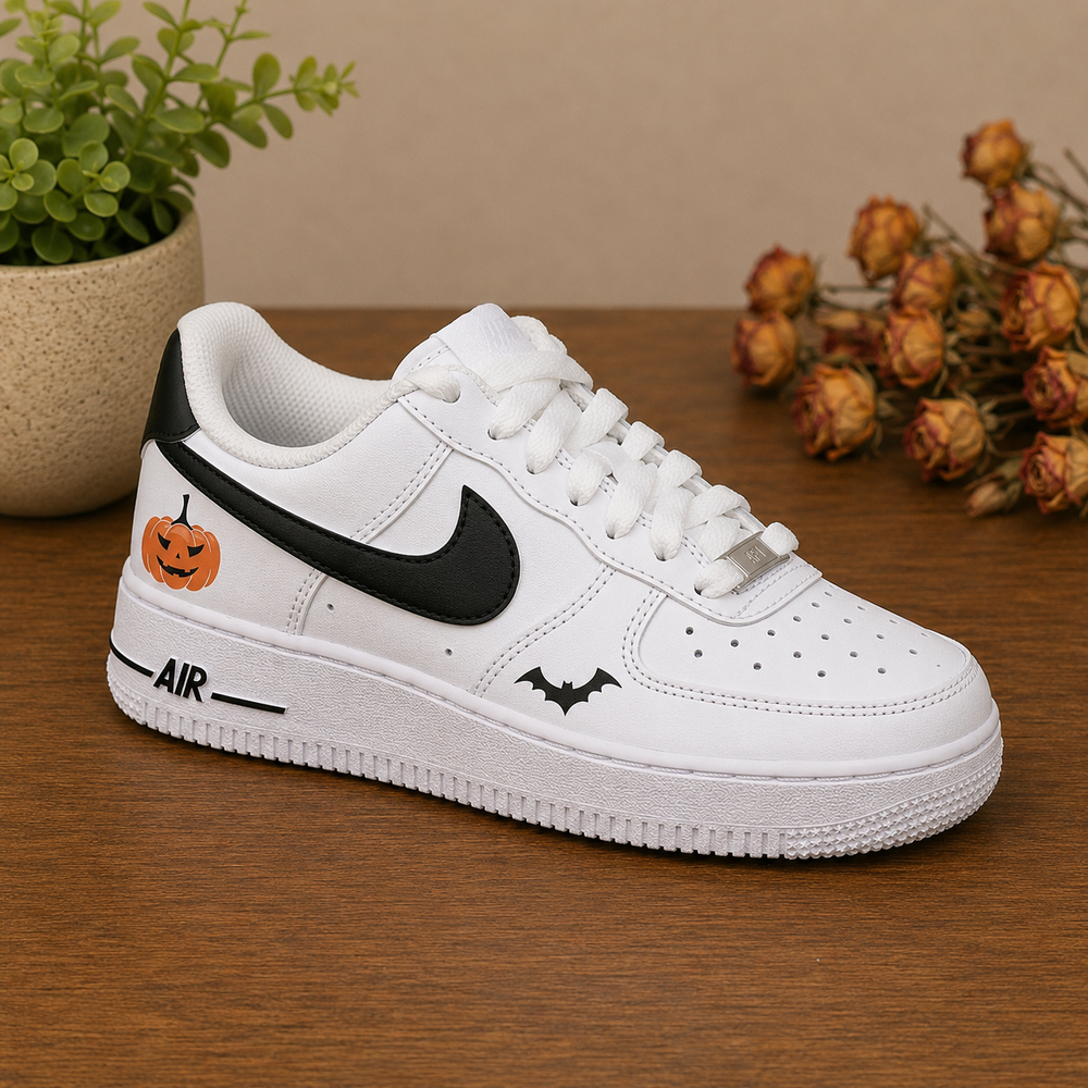 A white low-top sneaker with a black Swoosh, a black bat graphic, and an orange jack-o'-lantern graphic on the heel, displayed on a wooden surface with a potted plant and dried flowers in the background.