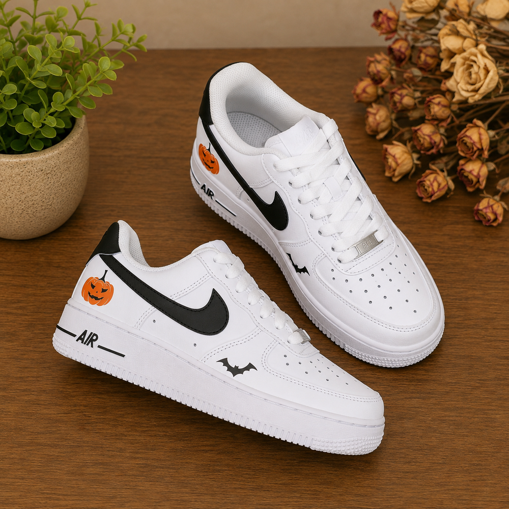 A pair of white Nike Air Force 1 style sneakers with black swooshes and Halloween-themed orange pumpkin and bat graphics, displayed on a wooden tabletop with a small potted plant and dried flowers in the background.