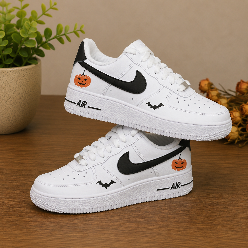 A pair of white Nike Air Force 1 Low sneakers with Halloween-themed graphics, including orange jack-o'-lanterns and black bats, displayed on a wooden surface with a potted plant in the background.