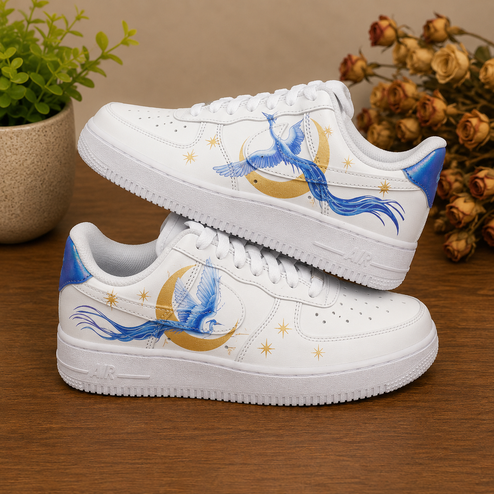 A pair of white low-top sneakers with blue phoenix and gold crescent moon designs, displayed on a wooden surface with decorative plants in the background.