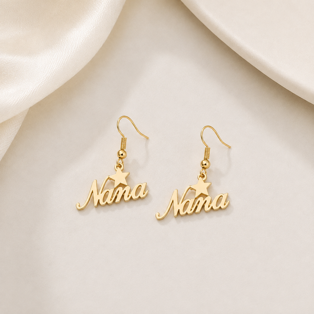 A pair of gold name earrings spelling 'Nana' with a small star charm above each name, displayed on a soft cream satin surface.