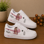 A pair of white Air Force 1 style sneakers with custom illustrations and text, displayed on a wooden surface with a potted plant and dried flowers in the background.