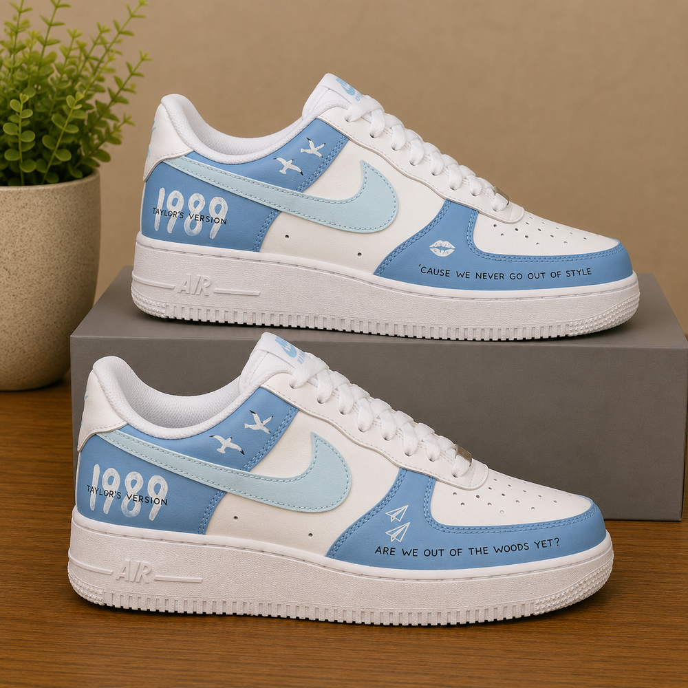 A pair of Nike Air Force 1 style sneakers in white, light blue, and medium blue, displayed on a gray shoebox on a wooden table with a potted plant in the background.
