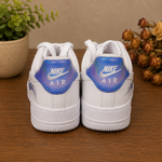 A pair of white Nike Air Force 1 Low sneakers shown from the back, featuring iridescent blue and purple heel tabs with the white Nike Air logo. The shoes are displayed on a wooden tabletop with a potted plant on the left and dried flowers on the right.