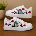 A pair of white low-top sneakers with custom Naruto character graphics, red cloud motifs, and black crows, displayed on a wooden surface with a potted plant in the background.