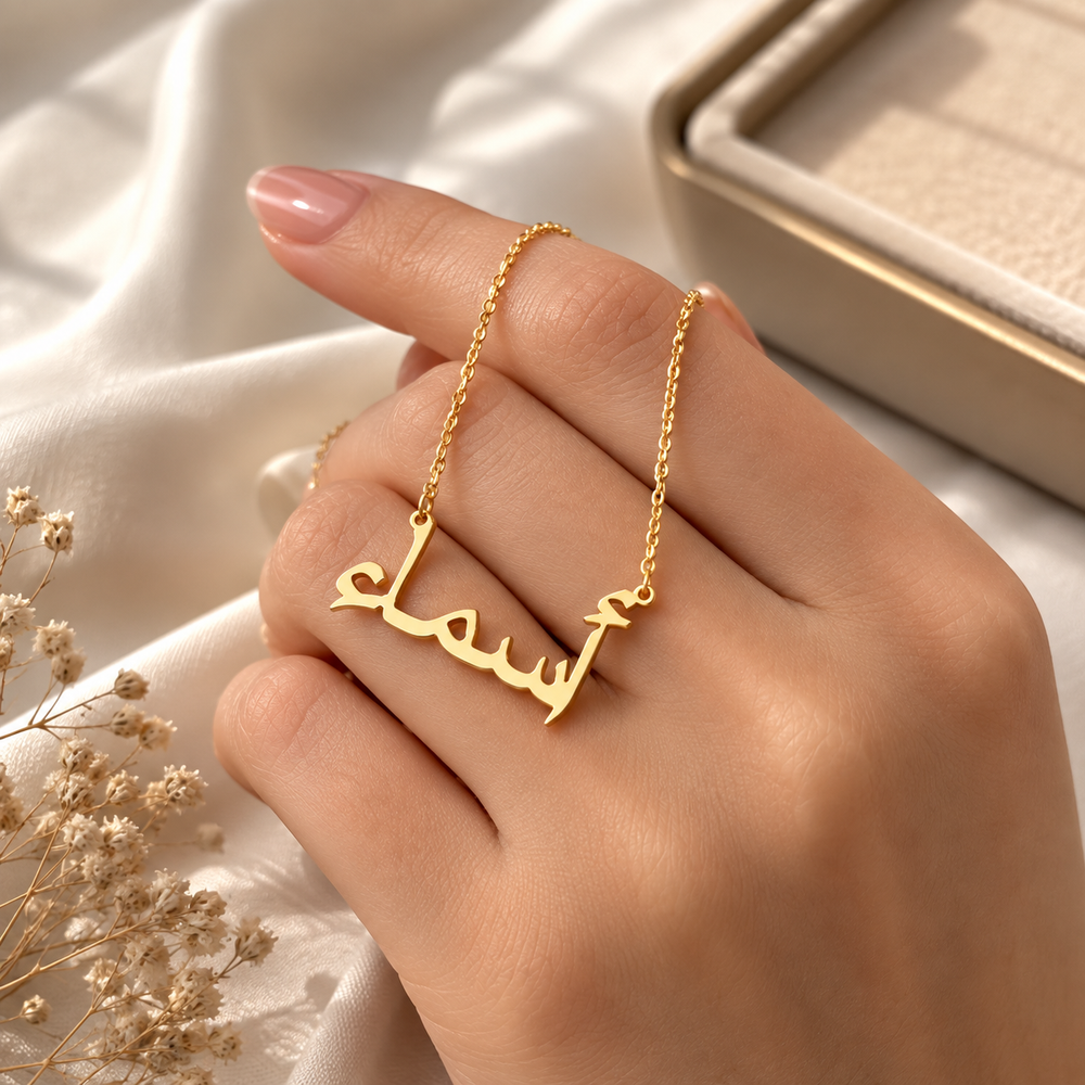 A gold nameplate necklace with Arabic script is shown worn on a person's finger, set against a soft white satin fabric background with a jewelry box in the distance.