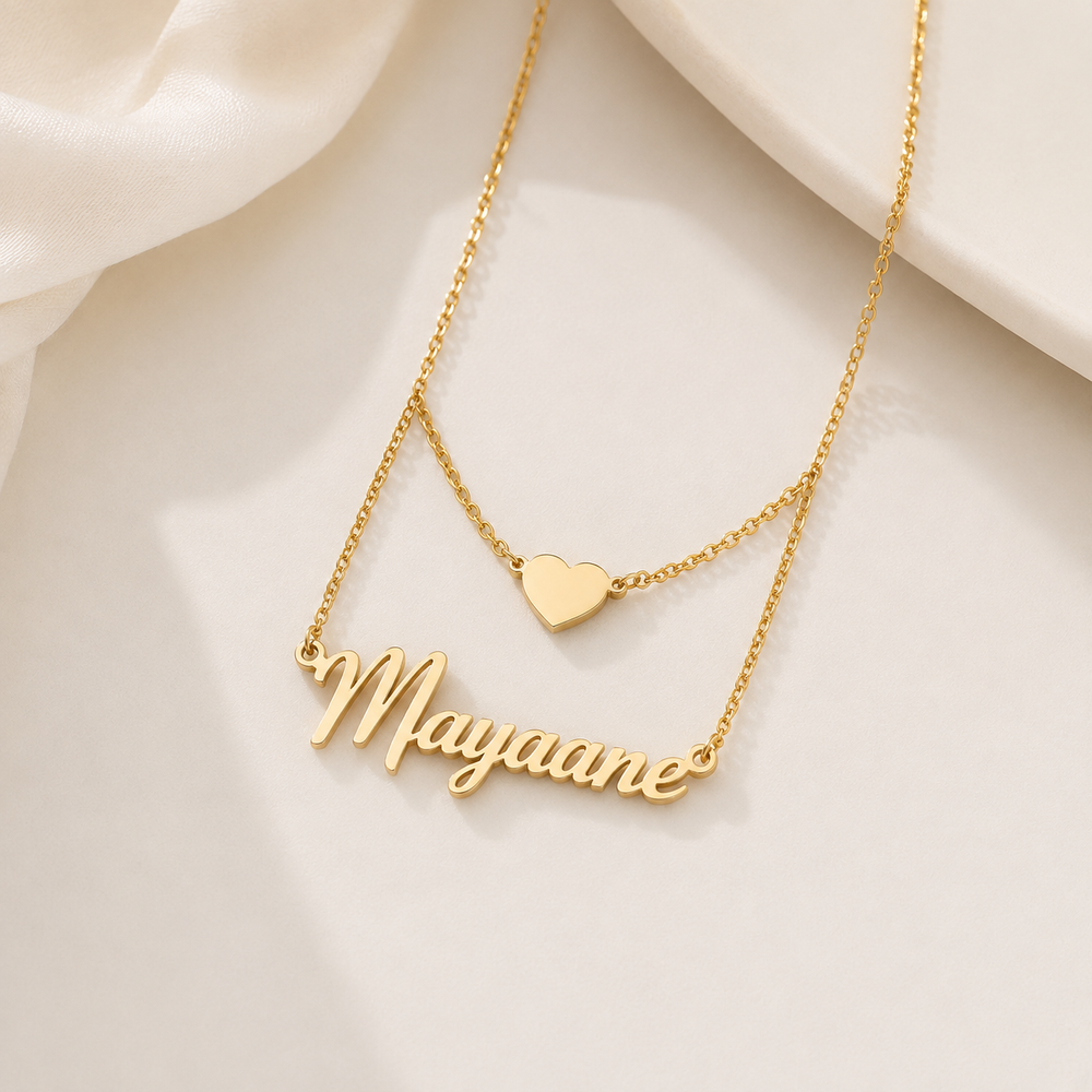A gold-tone layered necklace featuring a script nameplate reading 'Mayaane' and a small heart pendant, displayed on a soft cream fabric background.