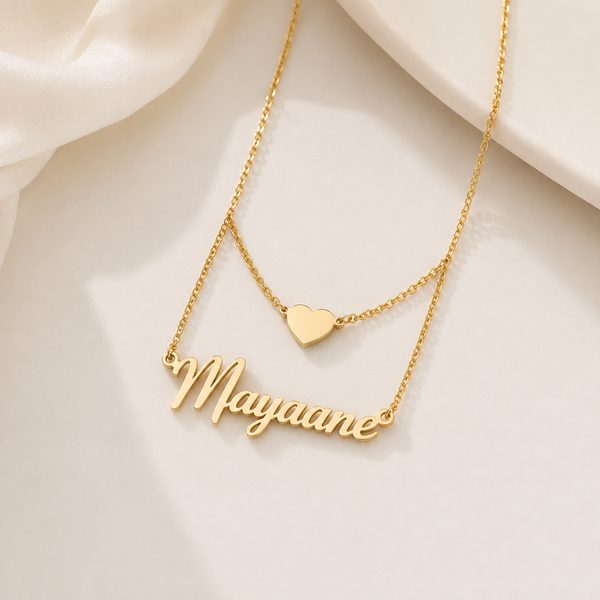 A gold-tone layered necklace featuring a script nameplate reading 'Mayaane' and a small heart pendant, displayed on a soft cream fabric background.