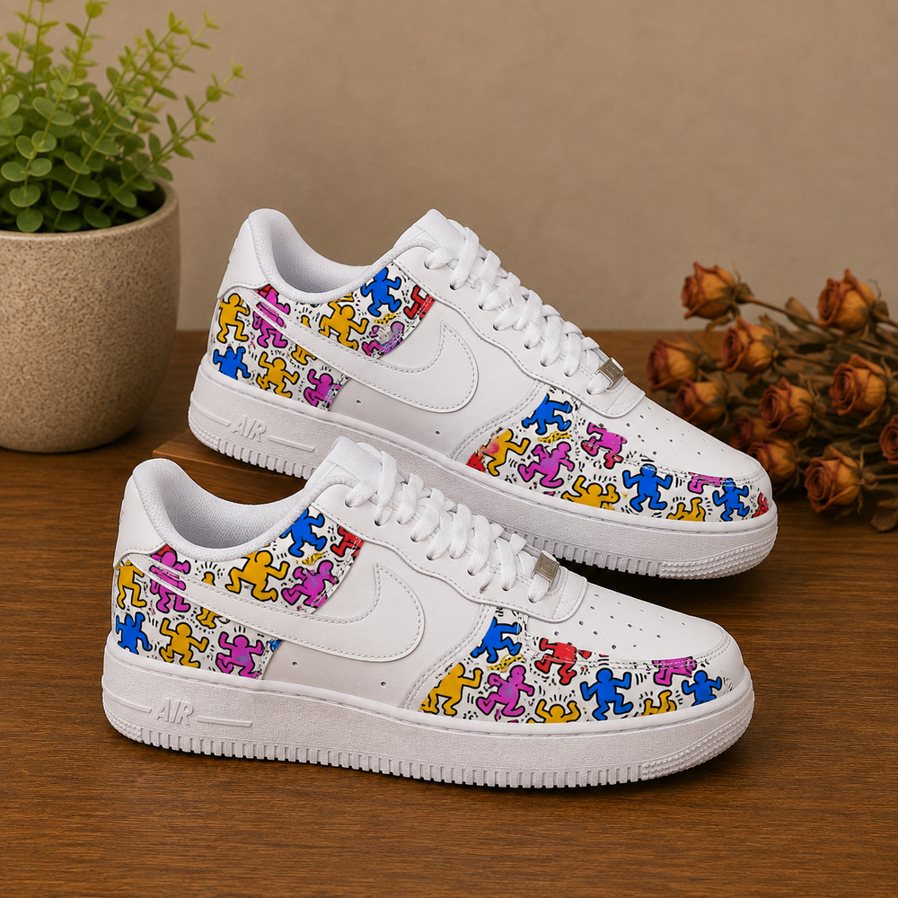 A pair of white Nike Air Force 1 Low sneakers decorated with colorful cartoon dancer patterns, displayed on a wooden table with a potted plant and dried flowers in the background.