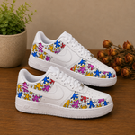 A pair of white Nike Air Force 1 Low sneakers decorated with colorful cartoon dancer patterns, displayed on a wooden table with a potted plant and dried flowers in the background.