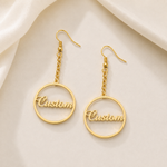 A pair of gold-tone custom name drop earrings with circular wire frames and the word 'Custom' in script, displayed on a soft cream satin background.
