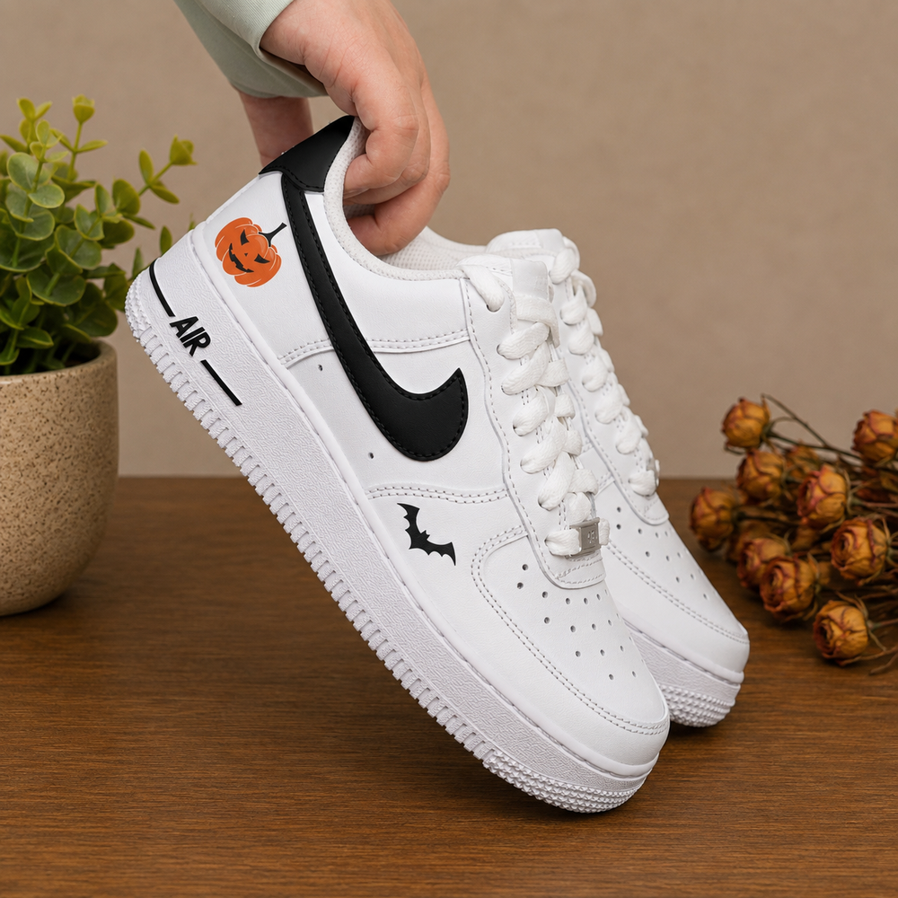 White Nike Air Force 1 sneakers with Halloween-themed graphics, including a black Swoosh, a bat silhouette, and an orange jack-o'-lantern on the heel, held by a hand over a wooden table with a potted plant and dried flowers.