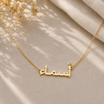A gold nameplate necklace with Arabic script lettering, displayed on a beige fabric background with soft shadows and dried white flowers in the corner.