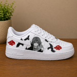 A white low-top sneaker with anime-style graphics, including a gray-haired character, black birds, and red cloud motifs, displayed on a wooden surface against a beige background.