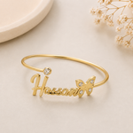 Gold nameplate cuff bracelet spelling 'Hassan' with a butterfly accent and a small clear stone, displayed on a light beige surface with dried flowers in the background.