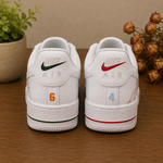 A pair of white Nike Air Force 1 low-top sneakers shown from the back, placed on a wooden surface with a potted plant on the left and dried flowers on the right. The left shoe has a green swoosh and orange number 6, while the right shoe has a red swoosh and light blue number 4.