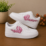A pair of white low-top sneakers with pink flame-like dragon illustrations on the sides, displayed on a wooden surface with a potted plant and dried flowers in the background.