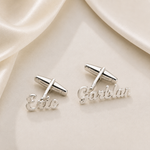 A pair of silver cufflinks displayed on a cream satin background. One cufflink features the name 'Eric' in cursive script, and the other features 'Gorblan' in cursive script.