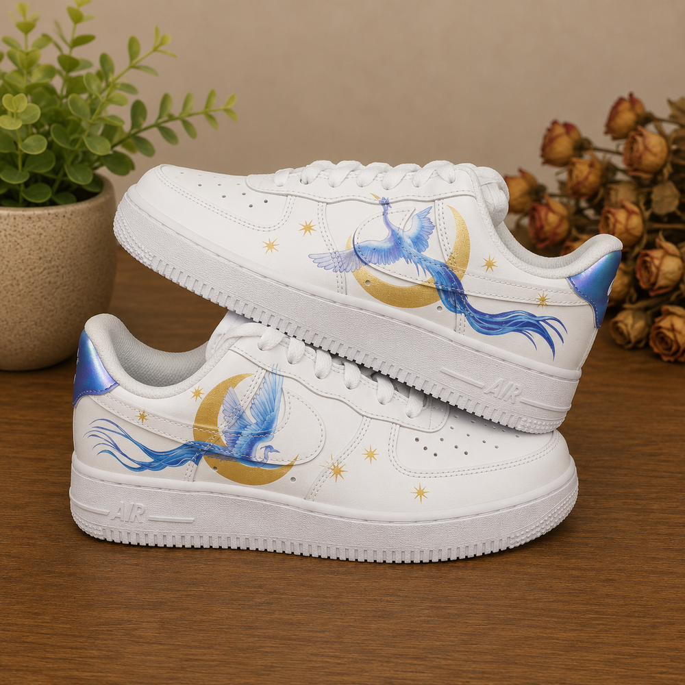 A pair of white low-top sneakers with custom blue phoenix and gold crescent moon designs, displayed on a wooden tabletop with decorative plants in the background.