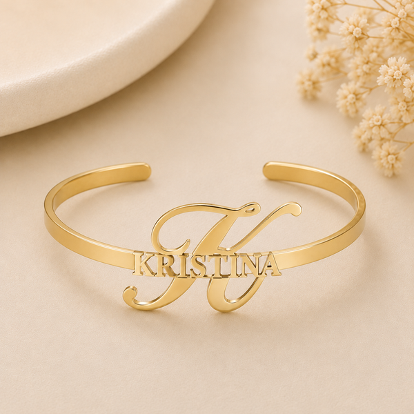 A polished gold-tone open cuff bracelet with the name 'KRISTINA' engraved across a large cursive letter 'K', displayed on a warm beige surface with dried flowers in the background.