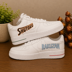 A pair of white low-top sneakers displayed on a wooden surface. The top shoe is positioned above the bottom shoe, with the top shoe's heel side showing the word 'SHUTOKU' in orange and black, and the bottom shoe's side panel showing 'RAKUZAN' in light blue.