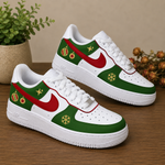 A pair of white Nike Air Force 1 style sneakers with green and red holiday-themed graphics, including ornaments, snowflakes, and stars, displayed on a wooden surface with a potted plant in the background.