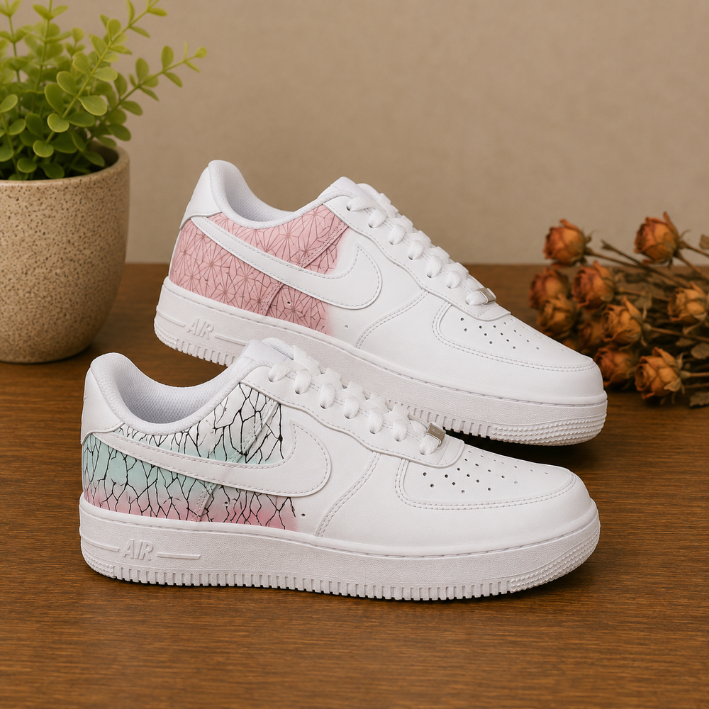 A pair of white low-top sneakers with pink and mint green cracked-pattern panels on the heel, displayed on a wooden surface with a potted plant and dried flowers in the background.