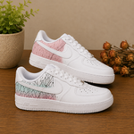 A pair of white low-top sneakers with pink and mint green cracked-pattern panels on the heel, displayed on a wooden surface with a potted plant and dried flowers in the background.